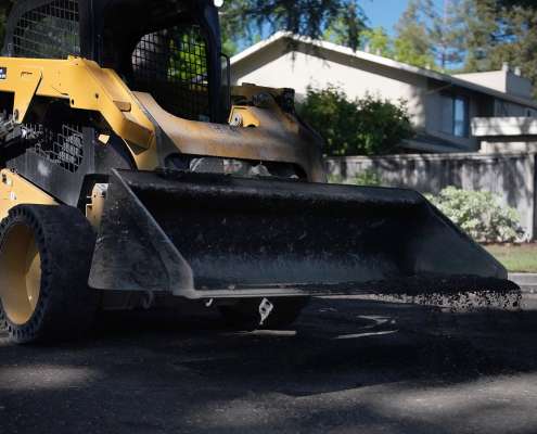 A Pacific Surfacing skid-steer spreads loose asphalt on a neighborhood street.