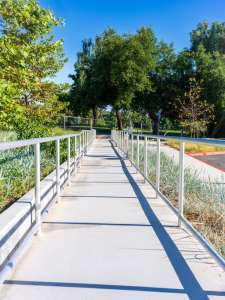 A concrete wheelchair-accessible ramp with metal railings on both sides, leading toward a park filled with green trees under a clear blue sky.